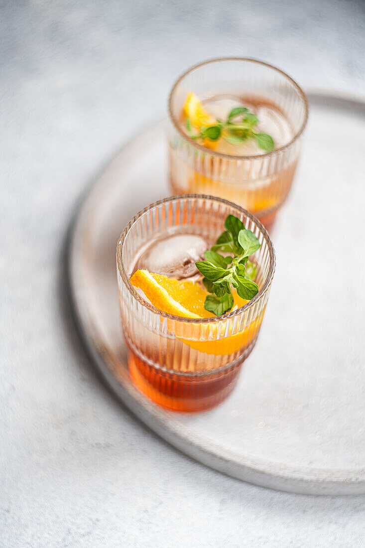 Zwei elegante Gläser mit einem lebhaften Sommercocktail aus Kirschlikör und Tonic, garniert mit einer frischen Orangenscheibe und Minzblättern, präsentiert auf einem schlichten, grauen Tablett