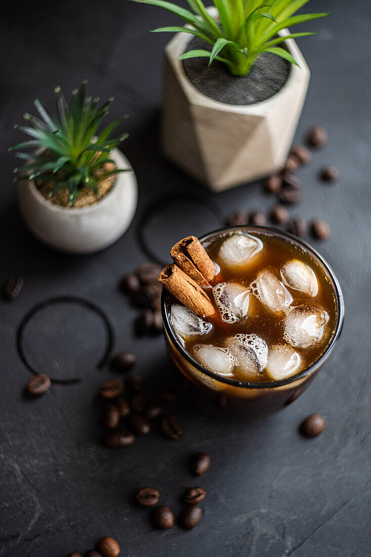 Draufsicht auf einen erfrischenden Eiskaffee, garniert mit Zimtstangen, umgeben von verstreuten Kaffeebohnen und Topfpflanzen auf einer dunklen Fläche, die eine gemütliche, aromatische Umgebung schafft.