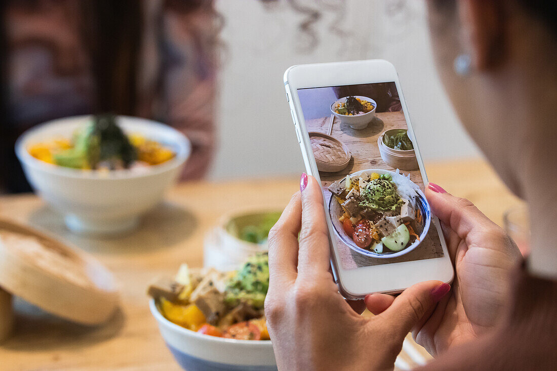 Ausgeschnittene, nicht erkennbare Frau, die mit ihrem Smartphone ein Foto von einem Poke-Gericht macht, während sie mit einer Freundin ein Wochenende im Restaurant verbringt