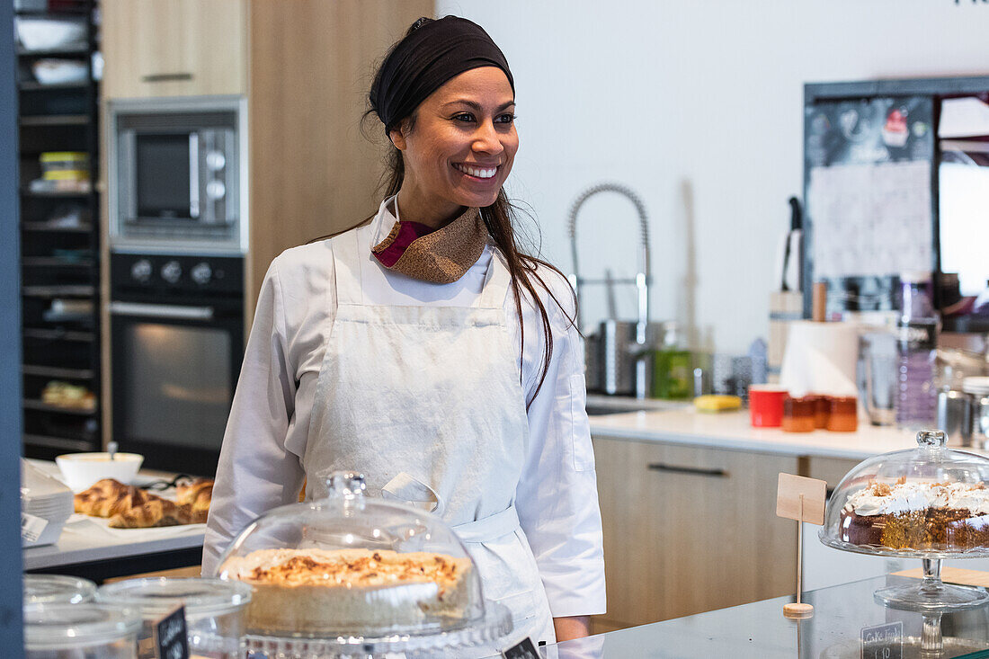 Positive Bäckerin in Uniform, die an der Theke mit veganen Desserts steht und wegschaut