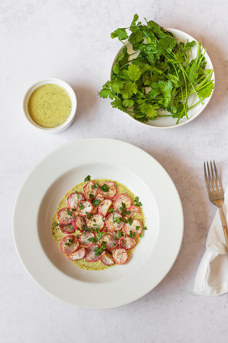 Top view of a dish with roasted radishes drizzled with green tahini sauce, fresh parsley on the side, and a bowl of sauce.