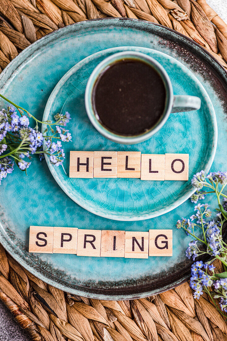 A cup of coffee and forget-me-not flowers on a turquoise plate with Hello Spring spelled out in letter tiles, evoking a fresh springtime mood