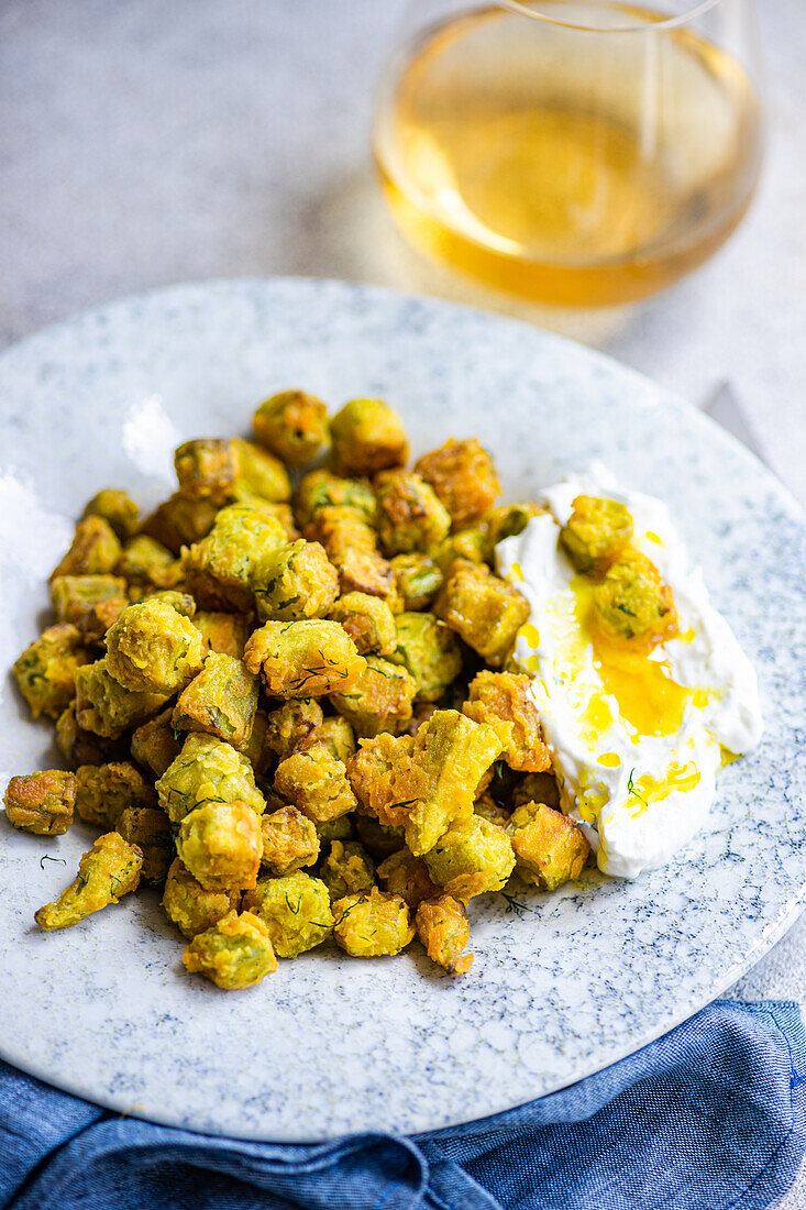 Knusprig gebratene Bamia, serviert mit einer cremigen, mit Dill und Knoblauch gewürzten Soße, dargestellt in einer rustikalen Umgebung mit einem erfrischenden Getränk im Hintergrund