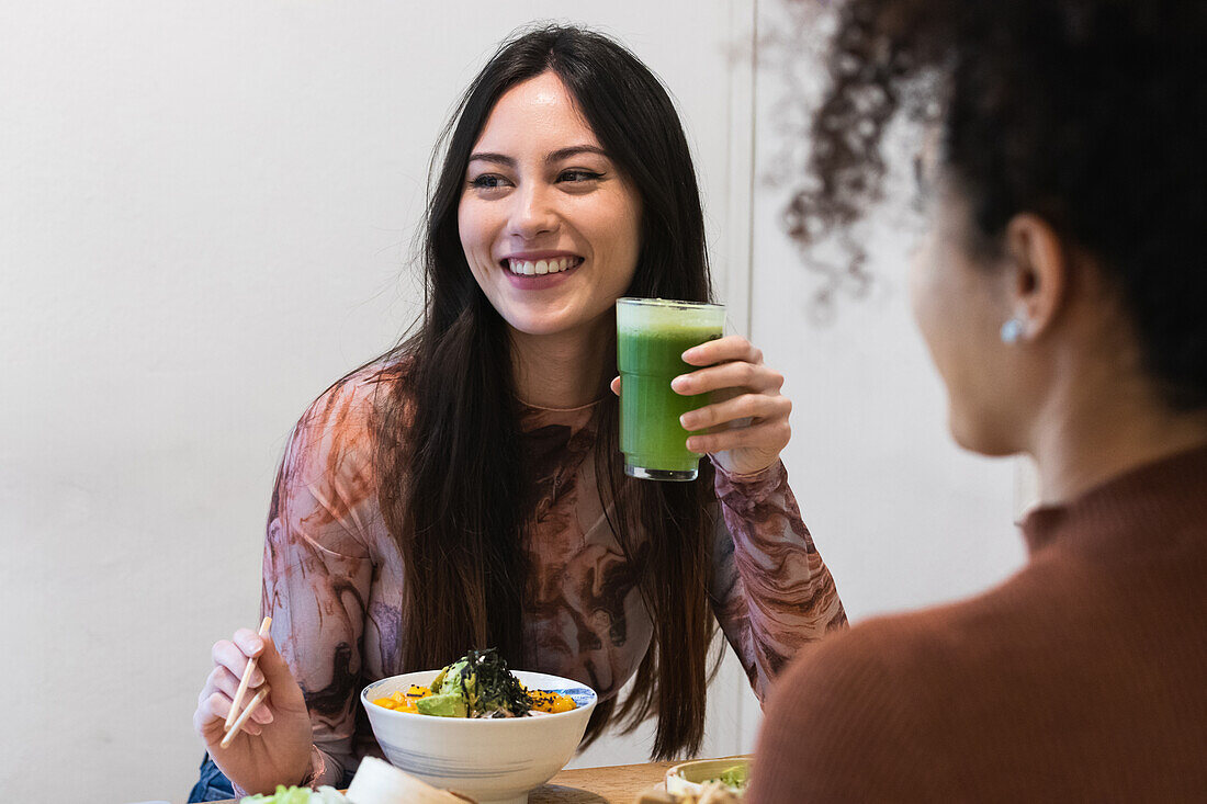 Zufriedene multiethnische Frau sitzt am Tisch und trinkt einen erfrischenden Smoothie, während sie mit einem nicht erkennbaren ethnischen Freund Poke isst