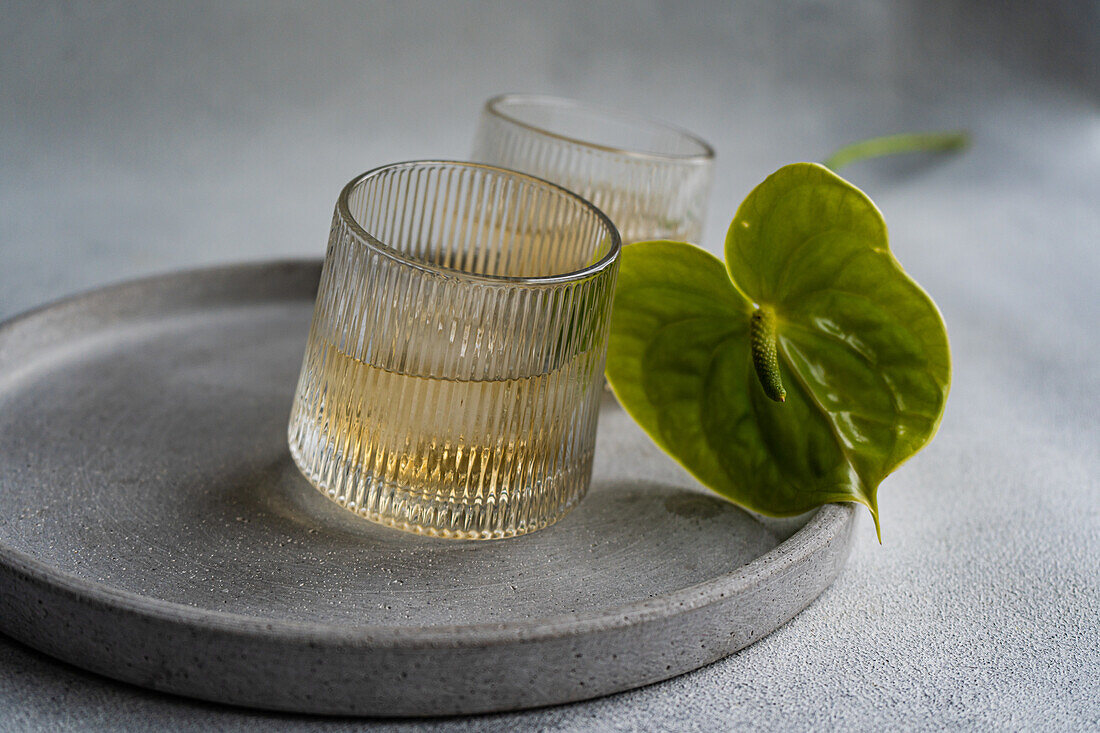 Elegante, minimalistische Tischdekoration mit einem Pfirsich-Wodka-Cocktail in einem strukturierten Glas neben einem leuchtenden Anthurium-Blatt, alles auf einer schlichten grauen Platte mit neutralem Hintergrund.