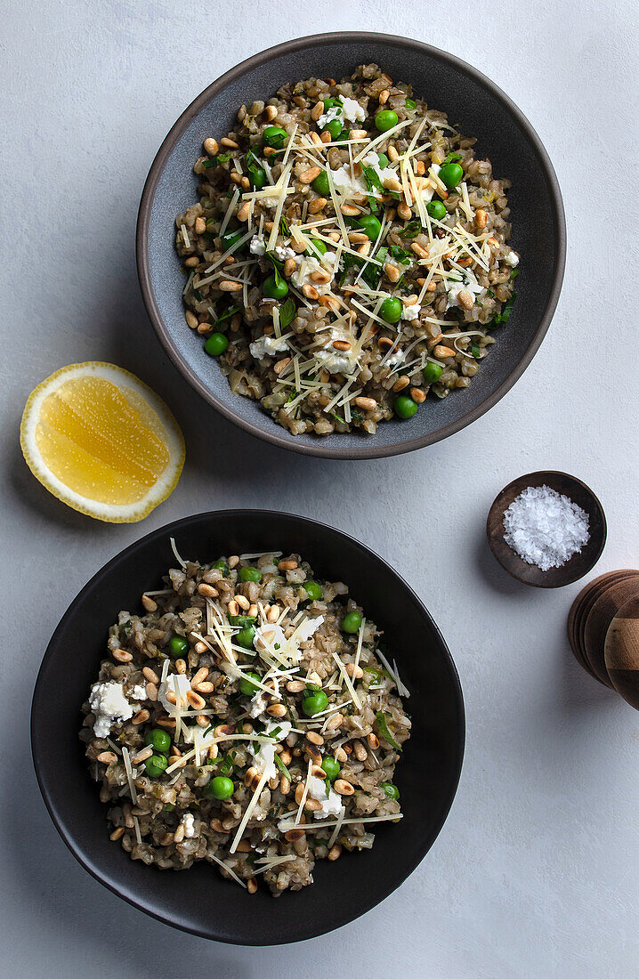 An overhead view of two bowls of barley risotto garnished with green peas and shaved cheese, alongside a lemon wedge and coarse salt.