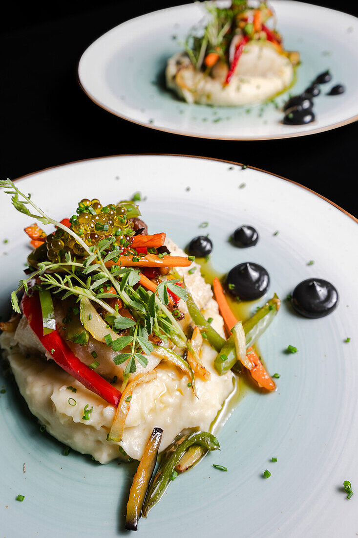 Elegant cod fillet served on a creamy mashed potato base, topped with colorful sauteed vegetables and a delicate herb garnish, presented on a ceramic plate.