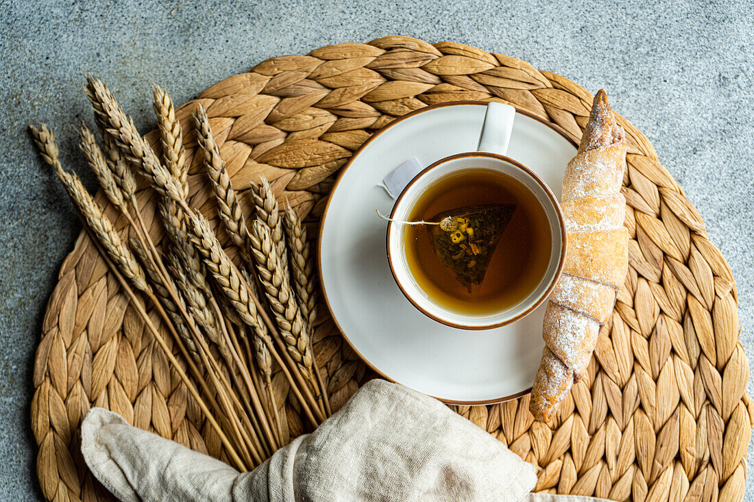 Top view of black tea with dried lemon slices in a white cup, paired with a sugar-dusted homemade croissant, placed on a woven placemat with wheat stalks.