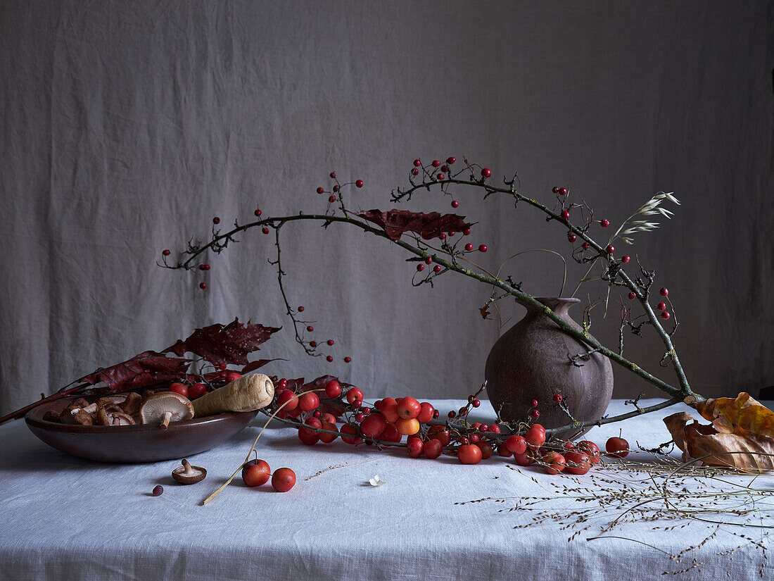 A moody and artistic still life arrangement featuring tiny red crab apples, shiitake mushrooms, and an intriguing vase. Set against a grey draped backdrop, this image captures the essence of autumn.