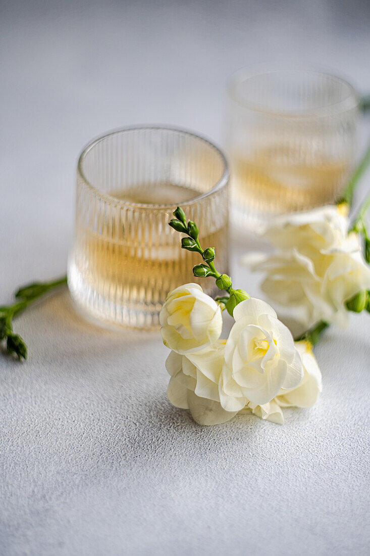 Elegante Pfirsich-Wodka-Cocktails, begleitet von blühenden weißen Freesien, präsentiert in strukturierten Gläsern auf einer ruhigen grauen Oberfläche.