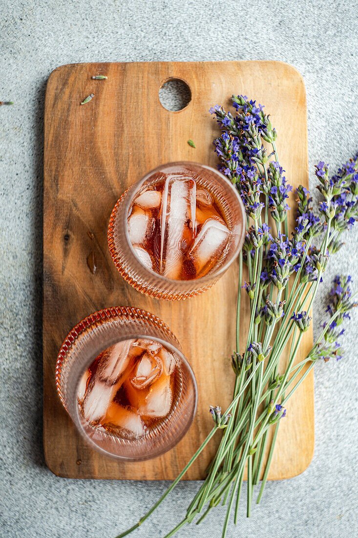 Zwei elegante Gläser mit Cognac-Cocktails, serviert auf einem rustikalen hölzernen Schneidebrett, begleitet von frischen Lavendelzweigen, die eine Mischung aus raffinierten Getränken mit einem blumigen Thema präsentieren