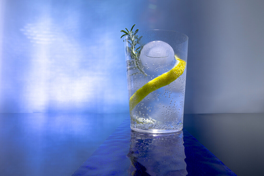 Refreshing gin tonic with ice sphere and lemon peel garnish on blue background