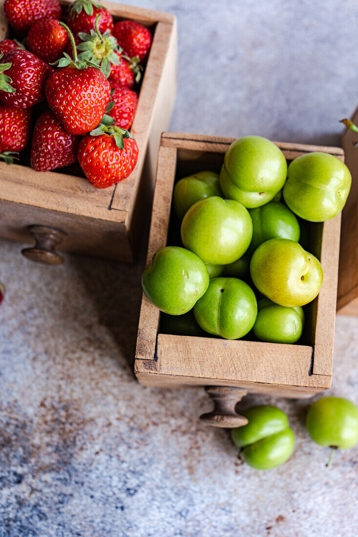 Reife Bio-Erdbeeren, Süßkirschen und grüne Pflaumen, arrangiert in alten Holzkisten auf einer strukturierten Oberfläche, die die farbenfrohe Fülle der Natur präsentiert