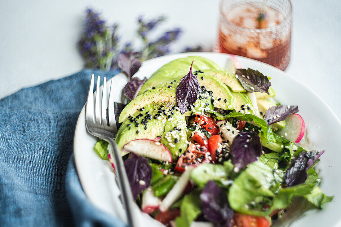 Bunter Gemüsesalat mit Kopfsalat, reifen Tomaten, knackigen Gurken, Radieschenscheiben, schwarzen Oliven und Avocado, bestreut mit schwarzem und weißem Sesam und roten Basilikumblättern. Serviert in einer weißen Schale mit einer Gabel an der Seite, perfekt für eine gesunde Mahlzeit.