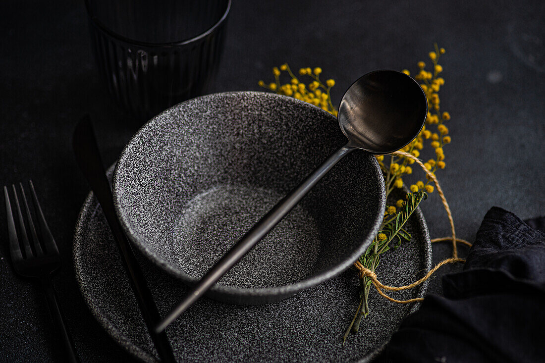 From above an artistic table setting featuring sleek black dinnerware, accented by vibrant yellow mimosa flowers and a touch of greenery.