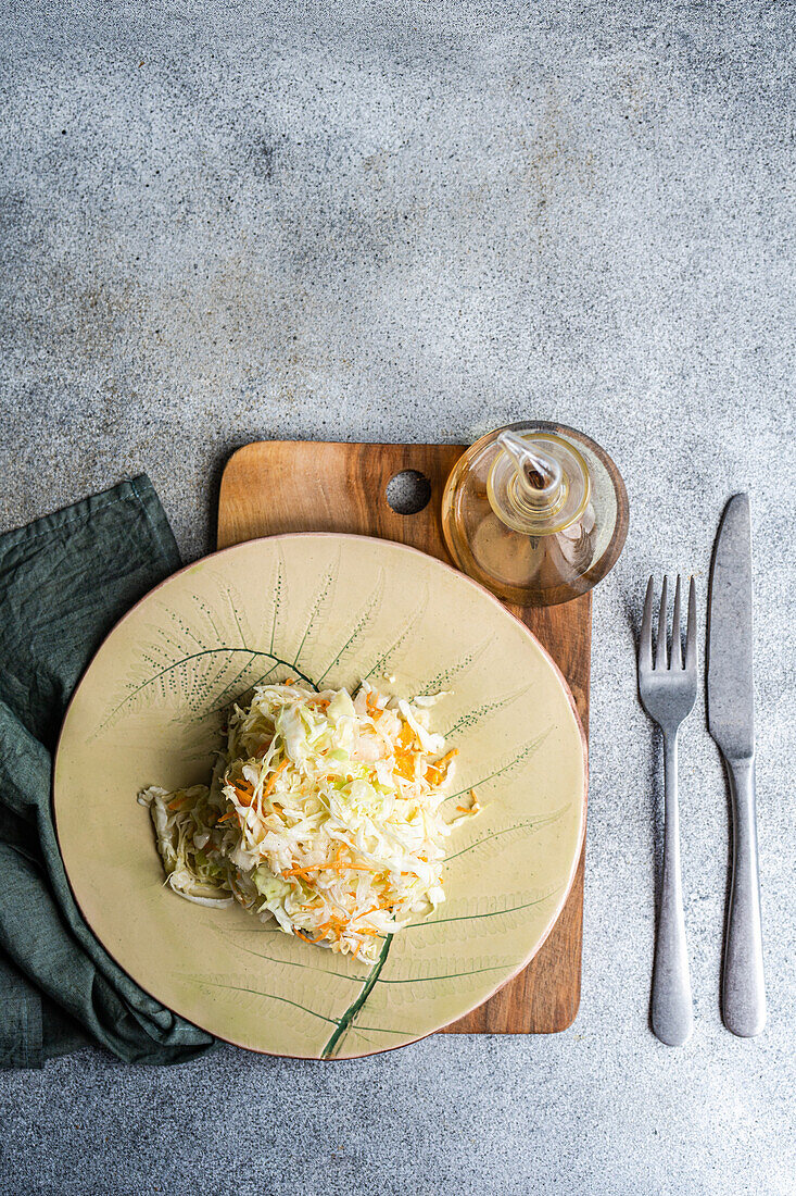 Top view of a nutritious organic cabbage and carrot salad drizzled with olive oil, presented on a ceramic plate, accompanied by a fork, knife, and a glass oil dispenser on a wooden cutting board.