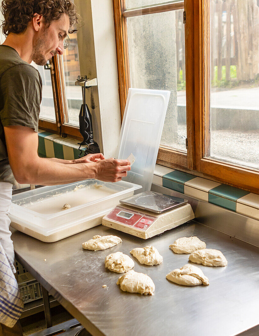 A baker meticulously weighs portions of pizza dough on a digital scale, ensuring consistent quality, amidst the surroundings of a natural lit, rustic kitchen