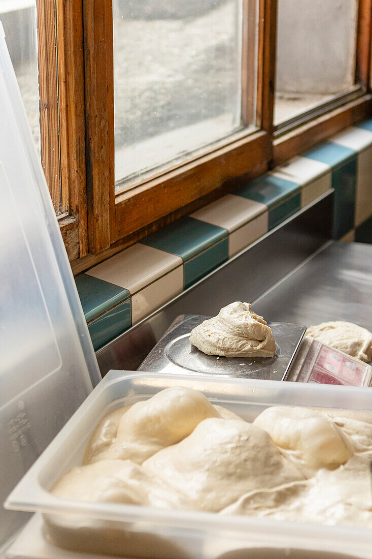 Das Bild zeigt frische Pizzateigkugeln, die auf einer Waage gären und in einem Plastikbehälter an einem sonnigen Fenster ruhen, und zeigt die ersten Schritte der Pizzazubereitung in einer rustikalen Küche