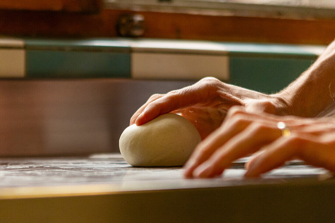 A pair of hands meticulously kneads fresh pizza dough on a marbled kitchen countertop, highlighted by warm, natural light