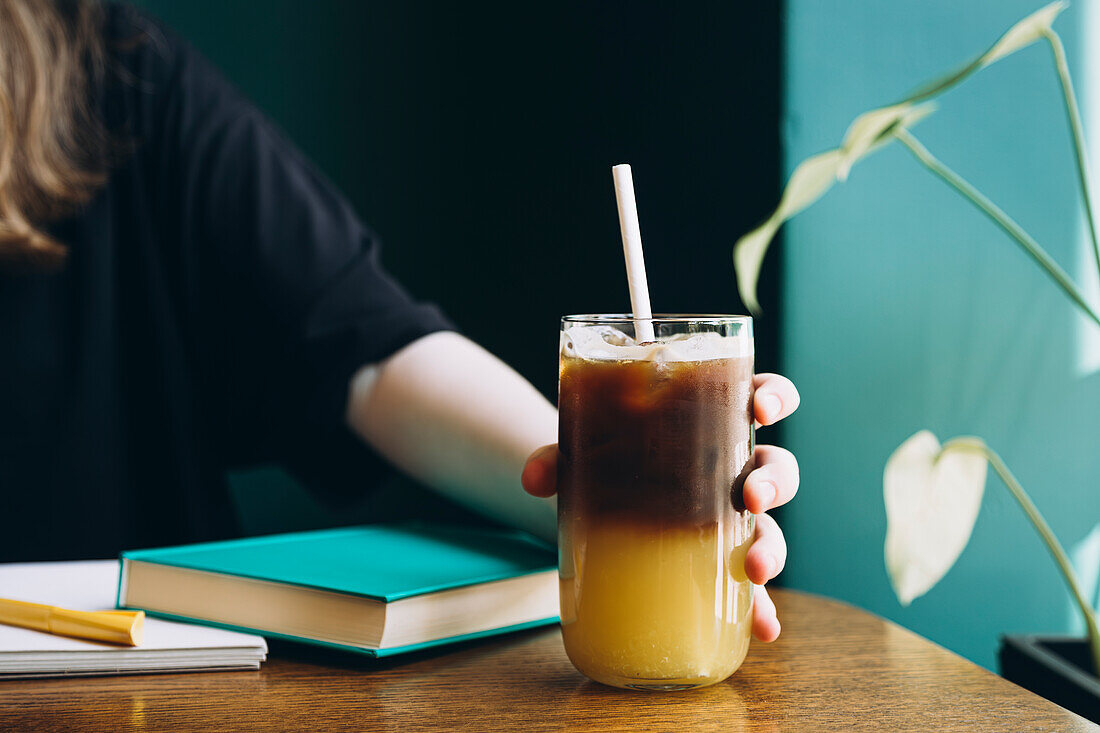 Abgeschnittene, nicht erkennbare Studentin mit einem gekühlten Kaffee in der Hand an einem Studientisch mit Büchern, die eine konzentrierte Lernumgebung zeigen.