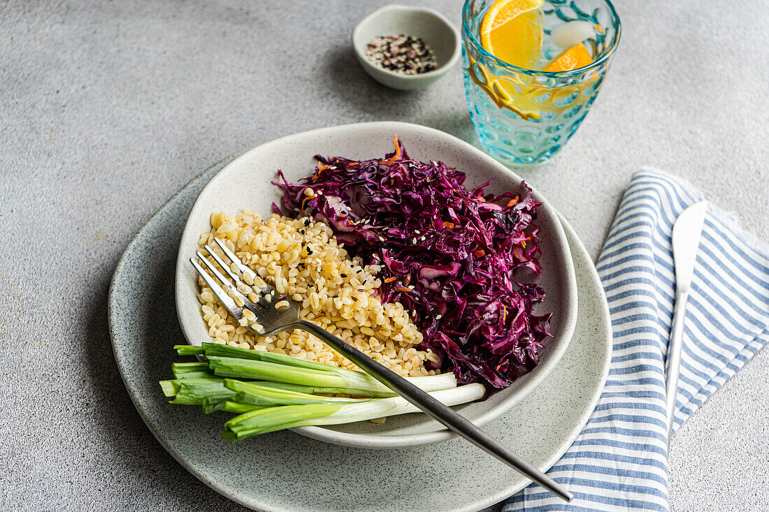 Eine nahrhafte Mahlzeit mit gekochtem Bulgur, gepaart mit einem leuchtenden Rotkohl- und Karottensalat auf einem hellgrauen Teller, ergänzt durch grüne Zwiebeln und ein erfrischendes Getränk