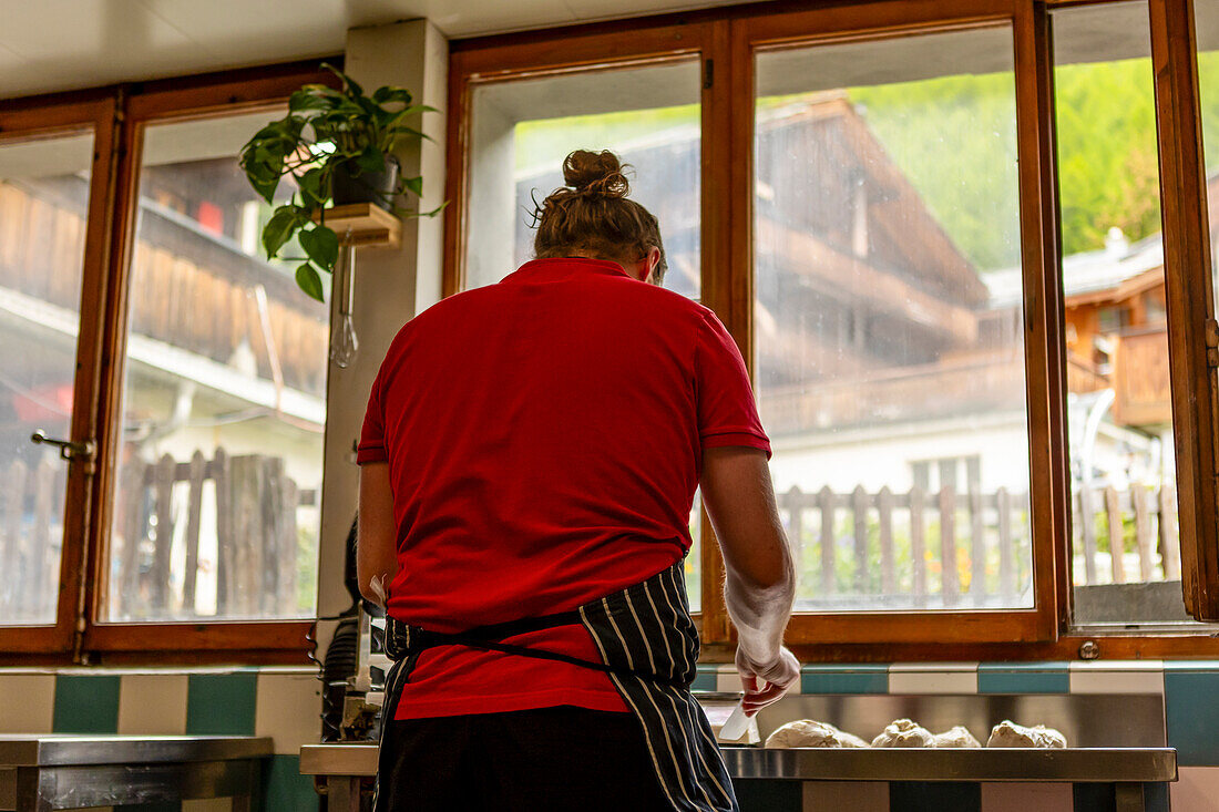 Ein Koch in rotem Hemd und gestreifter Schürze knetet Teig in einer gut beleuchteten Küche mit Blick auf ein Holzhaus vor dem Fenster