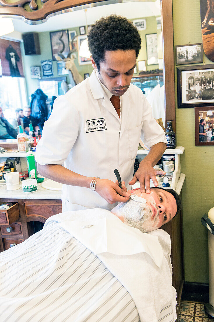 Professioneller Friseur und Haarschneider beim Rasieren seines Kunden im Schorem Barber Shop am Nieuwe Binnenweg, Rotterdam, Niederlande.