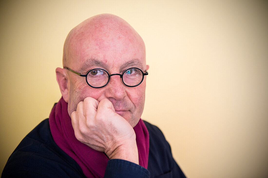 Tilburg, Netherlands. Portrait of bald, mature adult man wearing glasses, leaning in one hand against a yellow background.