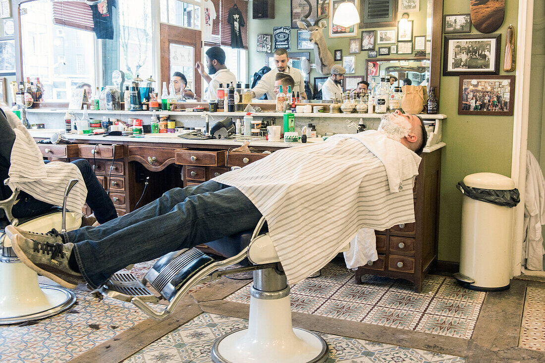 Junger Erwachsener, männlicher Kunde, der auf seine Nassrasur bei Schorem Barber Shop wartet. Nieuwe Binnenweg, Rotterdam, Niederlande.