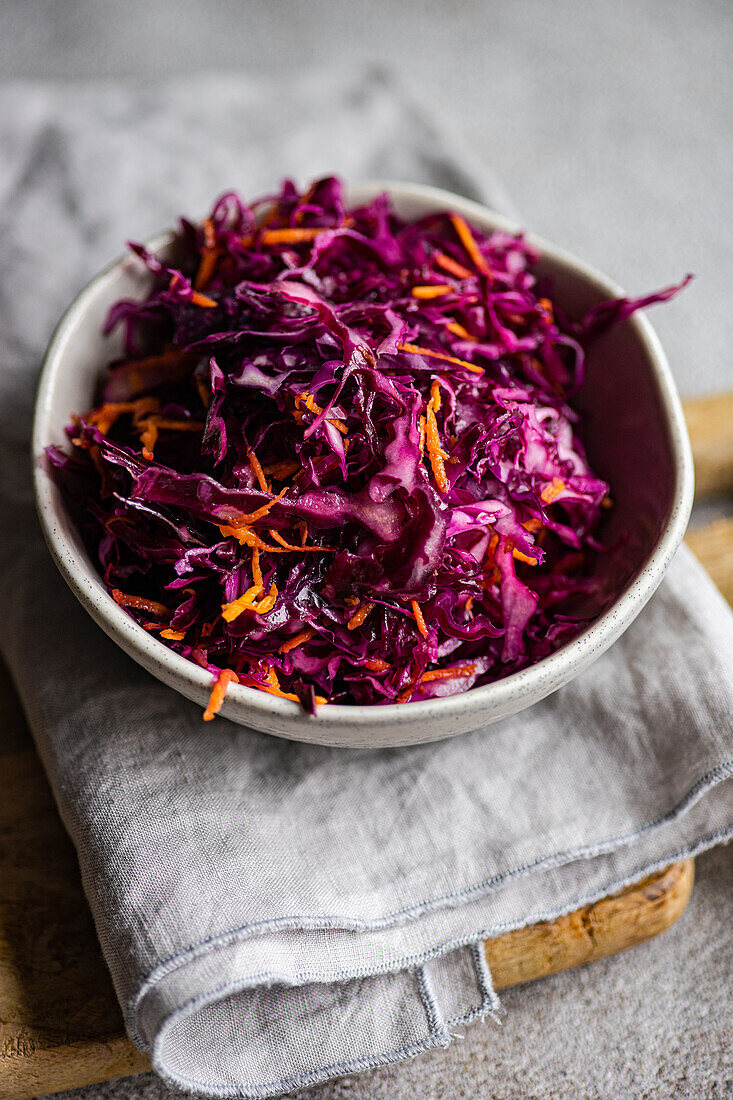 A healthy lunch set featuring a vibrant raw red cabbage and carrot salad paired with fluffy boiled bulgur, presented in a white bowl on a gray linen napkin