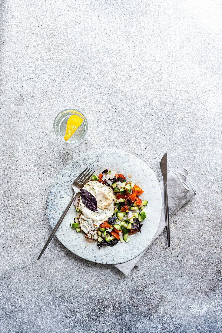 A nutritious keto-friendly lunch displayed on a marble patterned plate, featuring a perfectly egg atop a mix of colorful diced vegetables, accompanied by a slice of lemon