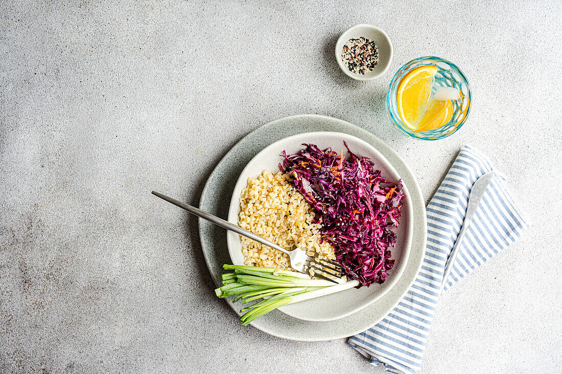 Ein nahrhaftes Mittagessen mit rohem Rotkohl- und Karottensalat und einer Portion Bulgur, begleitet von frischen grünen Zwiebeln, wird auf einem modernen grauen Teller mit gestreifter Serviette präsentiert