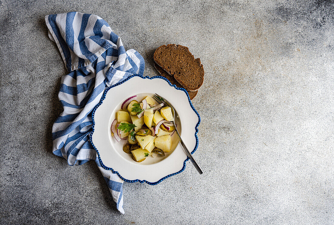 A fresh and appetizing German potato salad, consisting of pieces of boiled potatoes mixed with slices of red onion and fermented cucumber, garnished with parsley and served on a white plate with blue trim along with dark rye bread