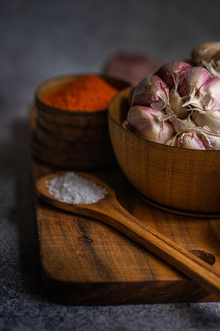 A low-lit close-up image showing a bulb of garlic, spices and salt arranged neatly on a wooden board, creating an atmospheric culinary theme