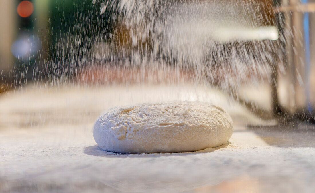 Dieses Bild zeigt eine Nahaufnahme einer Pizzateigkugel, die großzügig mit Mehl bestäubt wird. Die feinen Mehlpartikel schweben in der Luft und erzeugen eine dynamische und strukturierte Szene, die die ersten Schritte der Pizzaherstellung hervorhebt