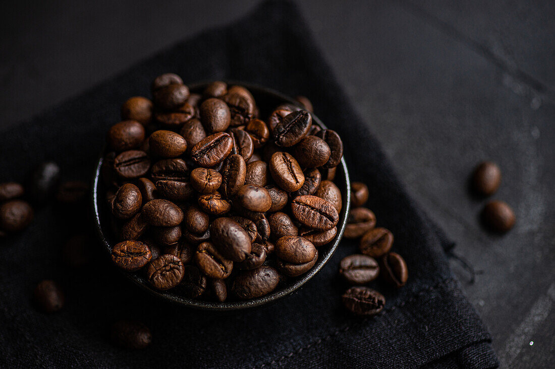 Draufsicht auf eine dunkle Schale mit glänzenden, frischen Kaffeebohnen vor einem dunkel strukturierten Hintergrund, der die reichhaltigen Strukturen und tiefbraunen Farben hervorhebt.