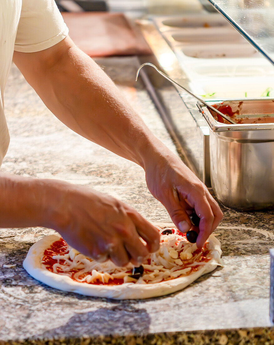 Man sieht die Hände einer Person, die einen Pizzaboden belegt. Die Umgebung ist eine gut ausgestattete Küche mit Zutaten wie Käse, Soße und Oliven im Vordergrund