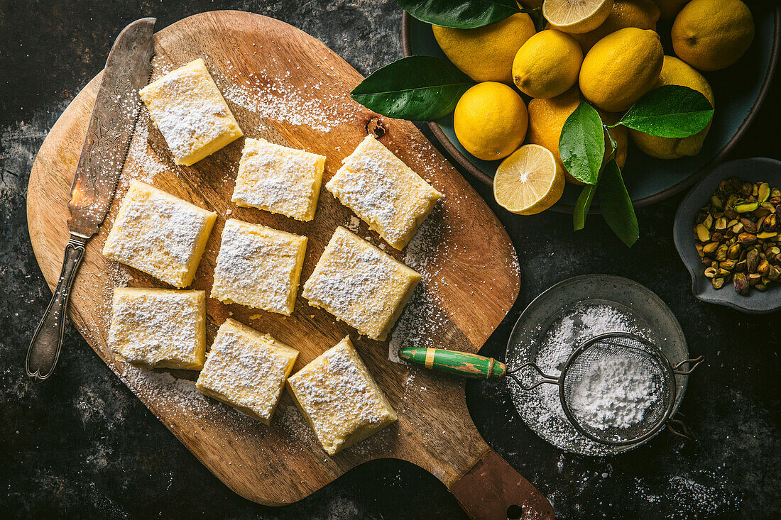 Lemon cake cut into squares on a wooden chopping board with a cake knife, icing sugar and a bowl of fresh lemons