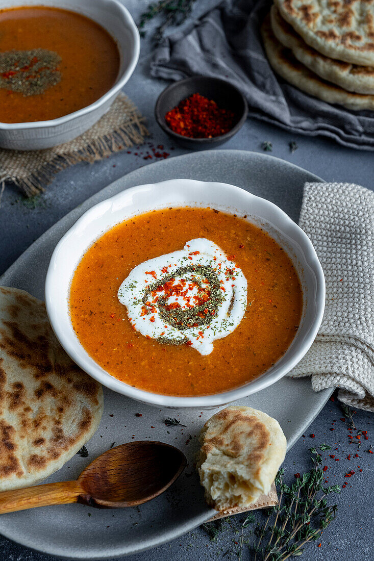 Tarhana soup with yoghurt, dried mint and red paprika flakes in a white bowl, with some flatbread and a wooden spoon.