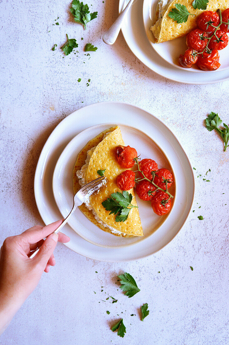 Glutenfreie Crepes mit Kräuter-Ricotta und gebratenen Rispentomaten. Weibliche Hand mit Gabel.