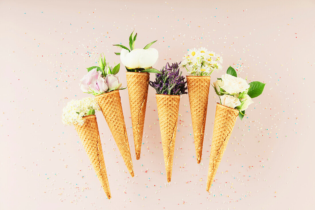 Flat-lay of waffle cones with flowers over pastel light pink background, top view. Spring or summer mood concept