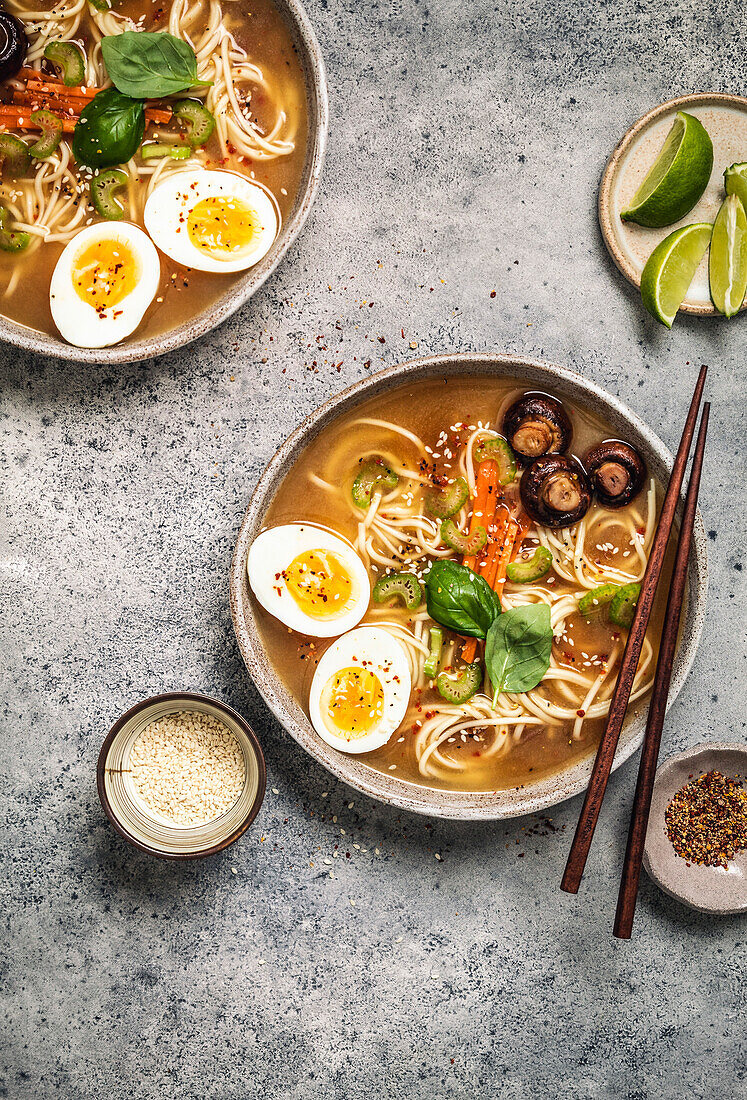 Ramen bowls with noodles, eggs, mushrooms, carrots and sesame seeds on a grey background