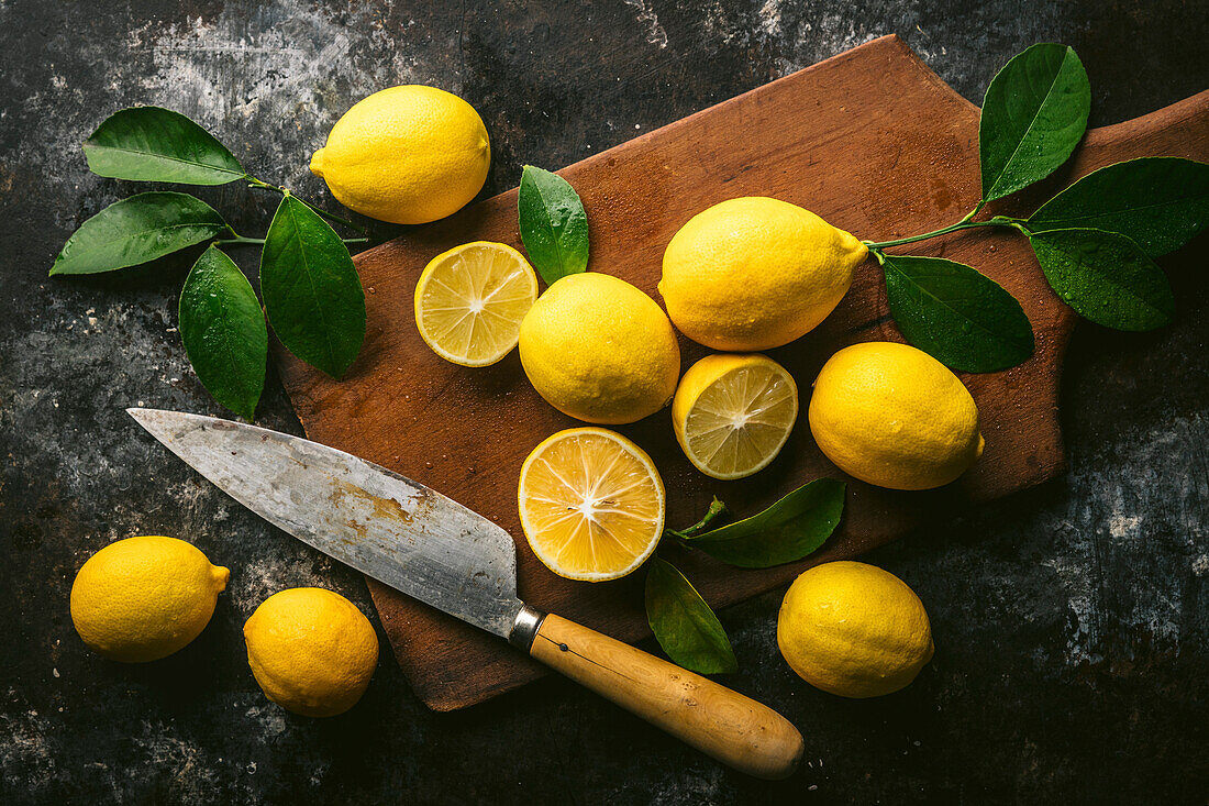 Freshly picked lemons with leaves, some halved, on an antique cutting board with a knife