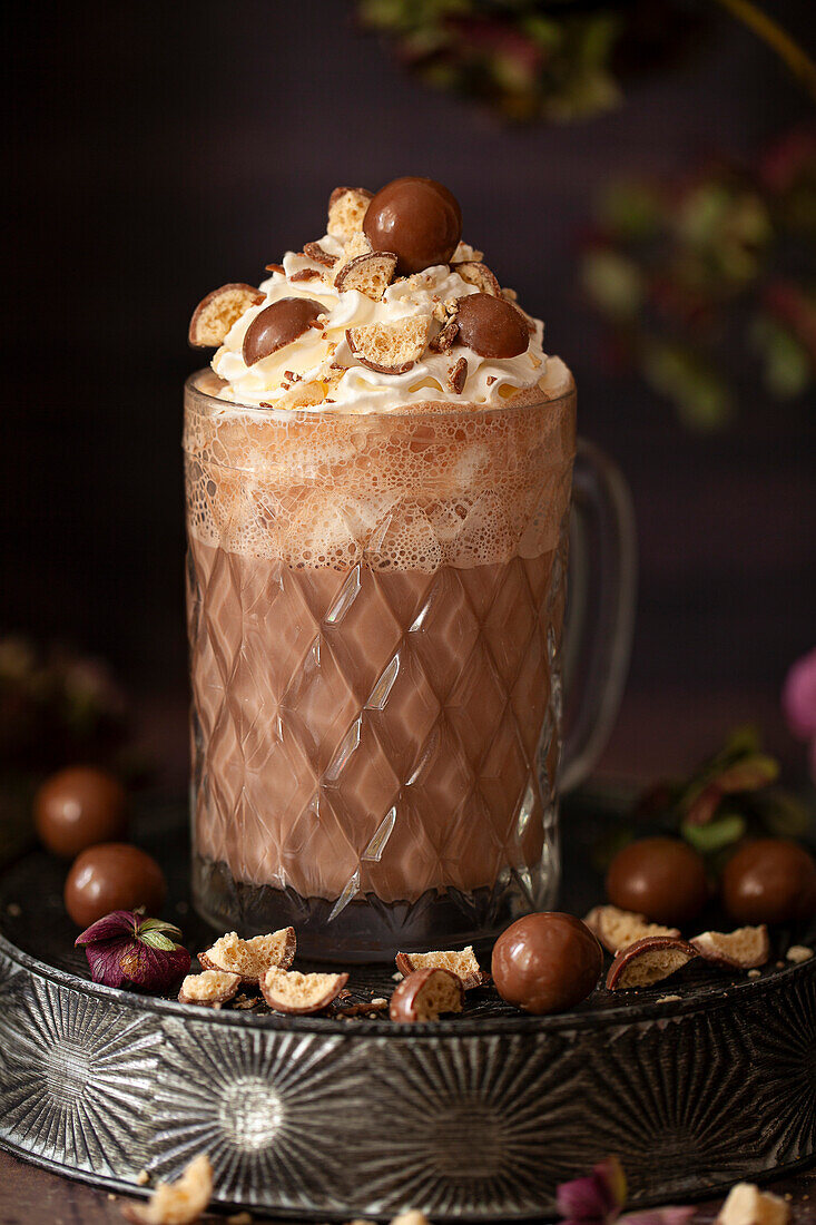 A hot chocolate in a vintage glass mug topped with whipped cream and honeycombe chocolate balls.