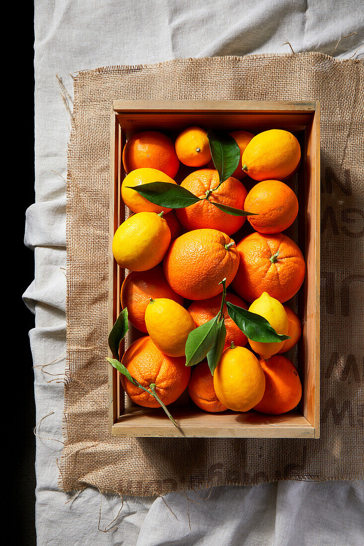 Navel-Orangen und Zitronen in einer Holzkiste auf einer Tischdecke aus Leinen und Sackleinen