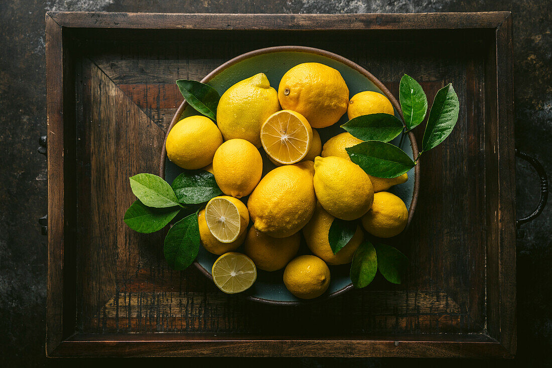 Assorted Lisbon and Meyer lemons, some cut, with fresh leaves, arranged in a blue ceramic bowl. In a rustic wooden crate