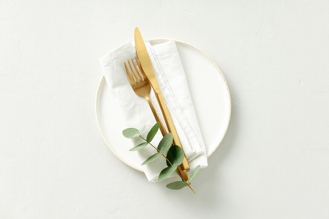 Gold Cutlery with eucalyptus branches on white plate with napkin over light grey Background. Minimalistic design. Copy Space