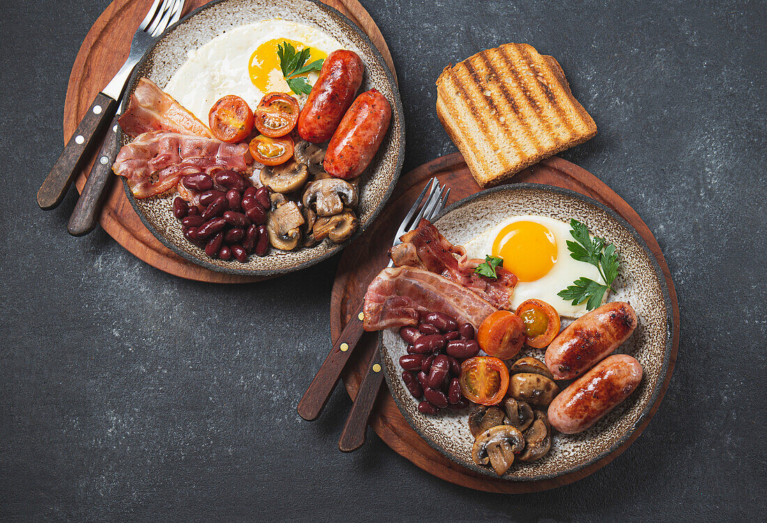 Traditionelles komplettes englisches Frühstück mit Spiegeleiern, Würstchen, Bohnen, Pilzen, gegrillten Tomaten, Speck und Toasts auf grauen Tellern