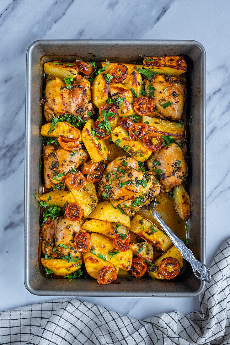 Baked chicken thighs, potatoes and cherry tomatoes garnished with parsley in a baking pan and a spoon in it photographed from top view.