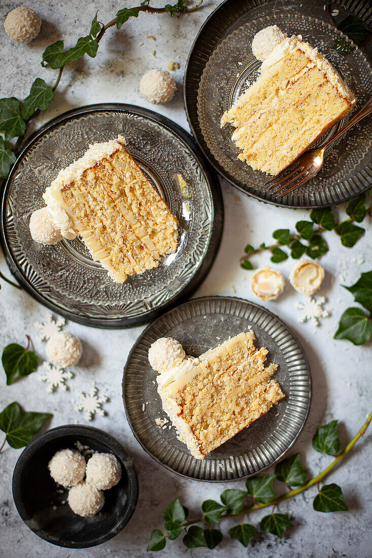 Drei Scheiben Torte aus weißer Schokolade und Kokosnuss auf Tellern mit Pralinen, die auf der Arbeitsfläche verstreut sind.